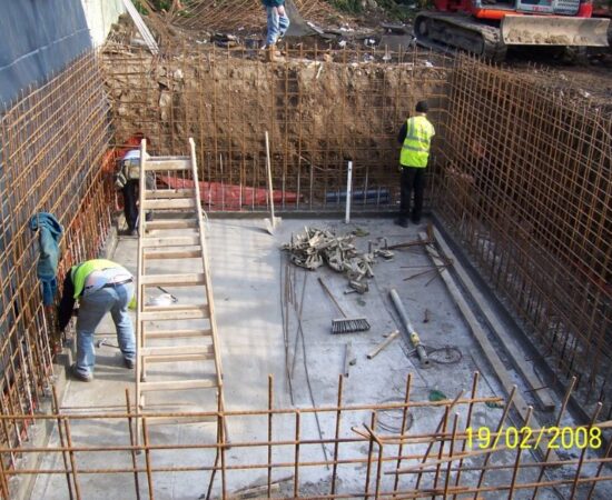 A photo showing three workmen in an approx. 10' deep hole with concrete floor and steel rebar in the walls (getting ready for the wall construction/pour).