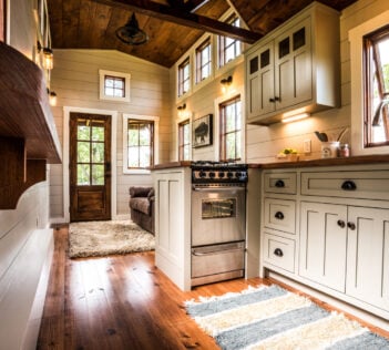 Timbercraft Tiny Homes' Denali gooseneck tiny house; interior of this home with light gray kitchen cabinets, loads of windows to bring in natural light and wood floors with rugs.