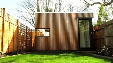 Backyard photograph showing this 'garden office pod', with vertical wood timber cladding and a second door into a storage area.